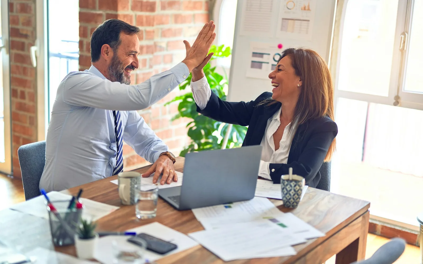 Two colleagues high-fiving across a work table covered in documents, with a laptop between them