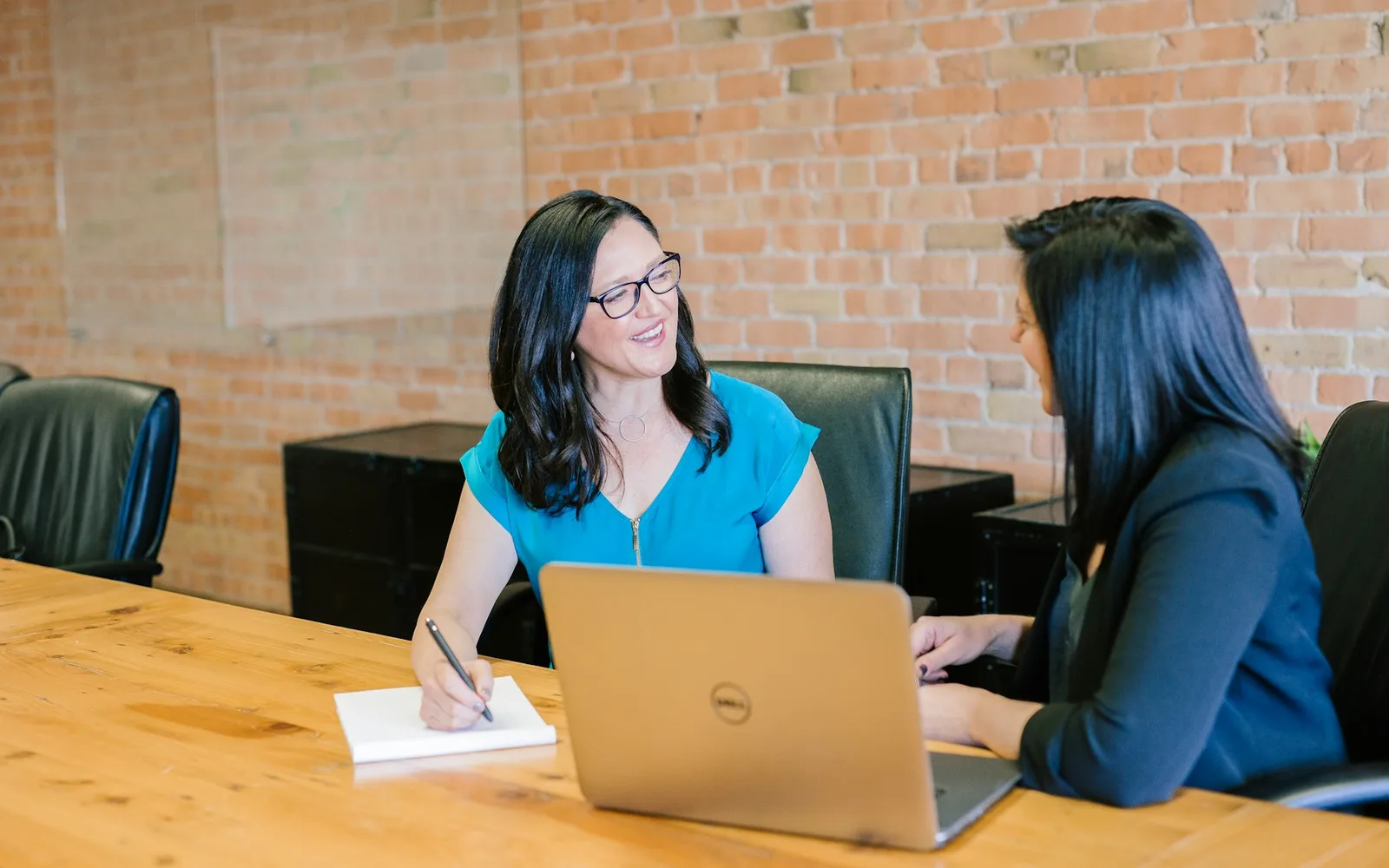 Two people in a consultation at a boardroom table, one taking notes beside a laptop